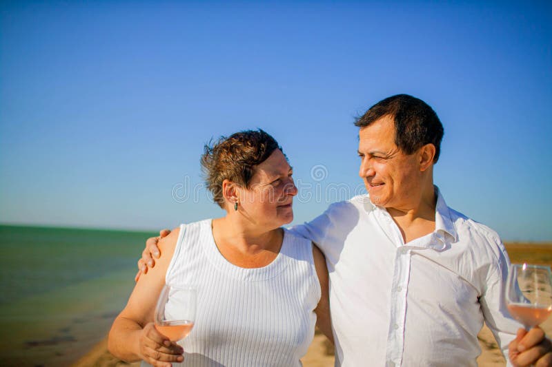 Porträtt från äldre par som står tillsammans med glas vin och omfamnar på stranden arkivfoto Porträtt från äldre par som står tillsammans med glas vin och omfamnar på stranden arkivfoto