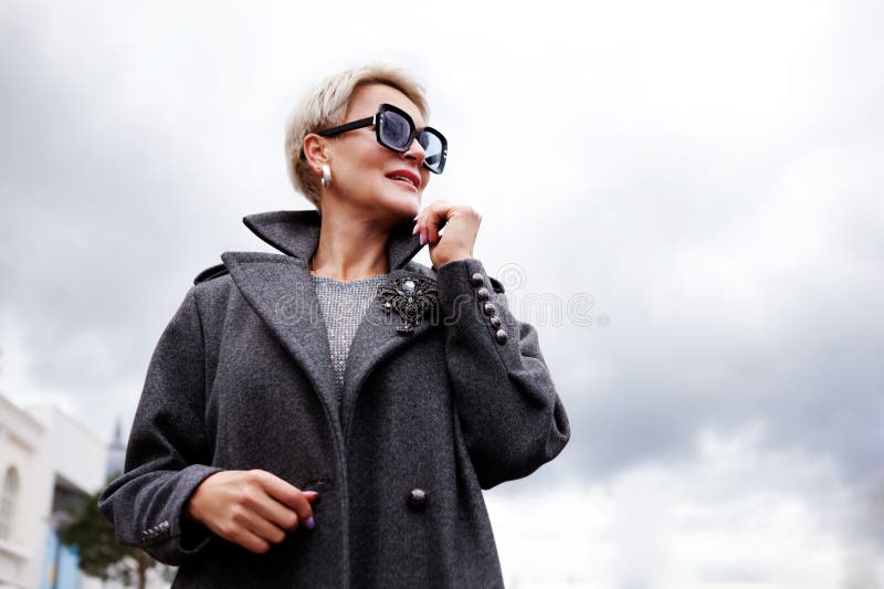 Portrait attractive female model on cloudy sky background. Modern stylish woman with short hair in gray double breasted pea coat and glasses. Portrait attractive female model on cloudy sky background. Modern stylish woman with short hair in gray double breasted pea coat and glasses.