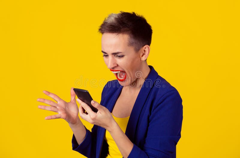 Portrait angry young woman screaming looking on mobile phone isolated on yellow wall background. Negative human emotions feelings. Short hair, blue suit, yellow shirt. Portrait angry young woman screaming looking on mobile phone isolated on yellow wall background. Negative human emotions feelings. Short hair, blue suit, yellow shirt