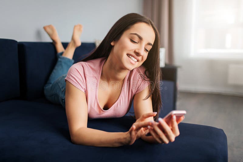Positieve jonge vrouw die op bank in ruimte liggen Zij raakt het scherm en het glimlachen van de telefoon Het model heeft rust royalty-vrije stock foto Positieve jonge vrouw die op bank in ruimte liggen Zij raakt het scherm en het glimlachen van de telefoon Het model heeft rust royalty-vrije stock foto