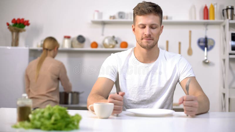 Positieve mens die op diner, jonge vrouwen kokende soep op achtergrond, honger wachten stock afbeeldingen Positieve mens die op diner, jonge vrouwen kokende soep op achtergrond, honger wachten stock afbeeldingen
