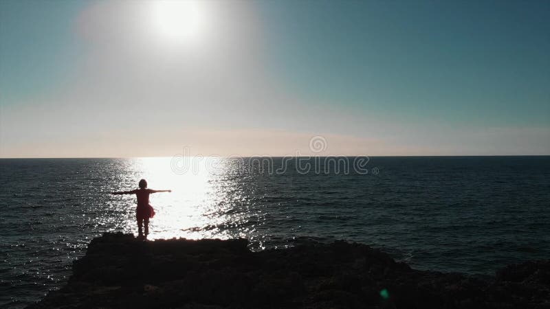 Position de silhouette de femme sur des falaises avec des mains à part en air contre le soleil et la route du soleil sur l'océan banque de vidéos Position de silhouette de femme sur des falaises avec des mains à part en air contre le soleil et la route du soleil sur l'océan banque de vidéos