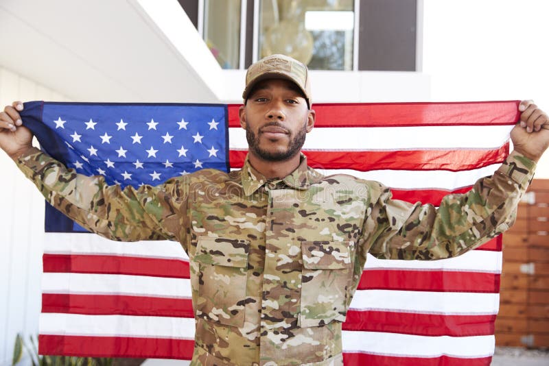 Position millénaire de soldat d'Afro-américain à l'extérieur du bâtiment moderne tenant le drapeau des USA, regardant à la caméra image libre de droits Position millénaire de soldat d'Afro-américain à l'extérieur du bâtiment moderne tenant le drapeau des USA, regardant à la caméra image libre de droits