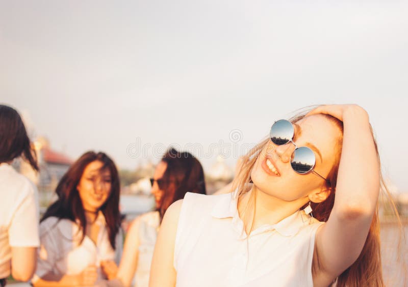 Positiv härlig lycklig röd haired flicka i spegelsolglasögon med vänner på bakgrund för blå himmel, sommarsolnedgångtid royaltyfria foton Positiv härlig lycklig röd haired flicka i spegelsolglasögon med vänner på bakgrund för blå himmel, sommarsolnedgångtid royaltyfria foton