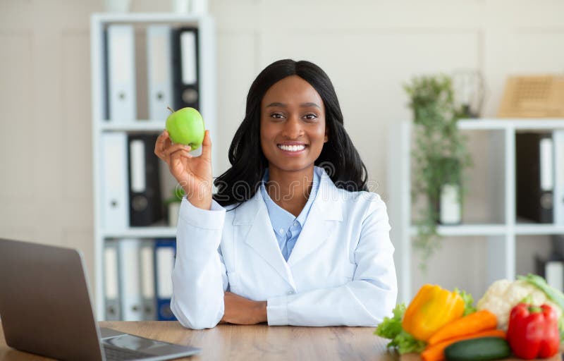 Positive black female dietitian holding apple fruit and looking at camera in clinic stock photos