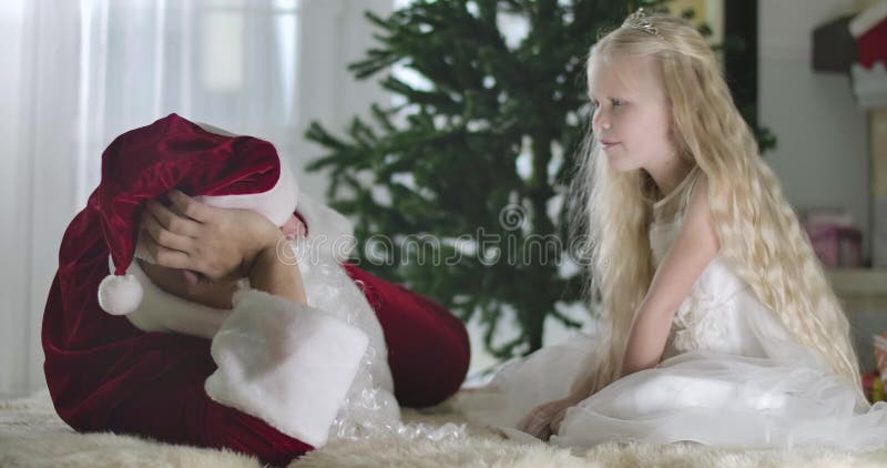 Positive Caucasian girl with blond curly hair making hush gesture and talking to Santa at home. Child spending Christmas