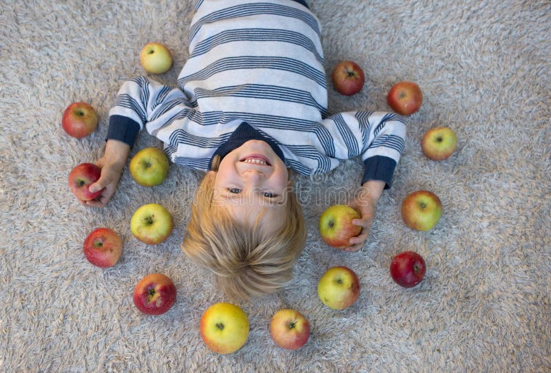 Positive cheerful boy of 4-5 years old in a striped sweater lies on his back, many apples are scattered around him