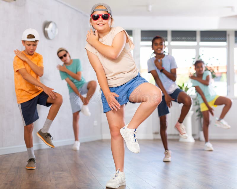 Positive female youngster in ball cap and dark glasses learns to dance, perform movements in choreographic class in company of peers. Young girls and guys repeat movements, train in spacious studio. Positive female youngster in ball cap and dark glasses learns to dance, perform movements in choreographic class in company of peers. Young girls and guys repeat movements, train in spacious studio