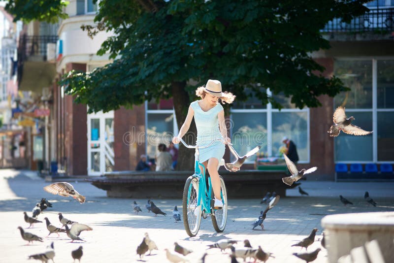 Positive girl in straw hat riding blue vintage bike in paved city center chasing pigeons flocks during hot summer day