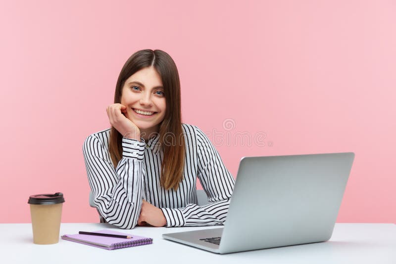 Positive inspired brunette woman sitting at laptop on her office and looking at camera with toothy smile, making notes and drinking coffee during work. Indoor studio shot isolated on pink background. Notes office stock images, royalty-free photos and pictures