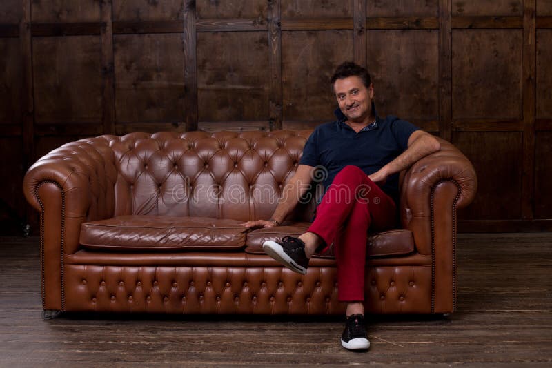 Positive middle aged man feeling glad and smiling while being in the laconic room and sitting on the blown sofa royalty free stock image Positive middle aged man feeling glad and smiling while being in the laconic room and sitting on the blown sofa royalty free stock image