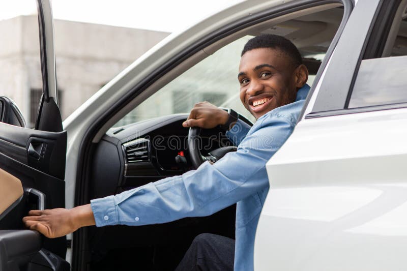 Positiver schwarzer Mann, der aus dem Auto rausgeht, öffnet Autotür und lächelt hinein. Auto-Testlaufbuchung stockbild
