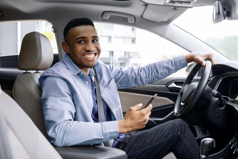 Positiver schwarzer Mann, der Smartphone benutzt, während er im modernen Auto sitzt und Kamera lächelt. Guy mit GPS-Navigator stockbild