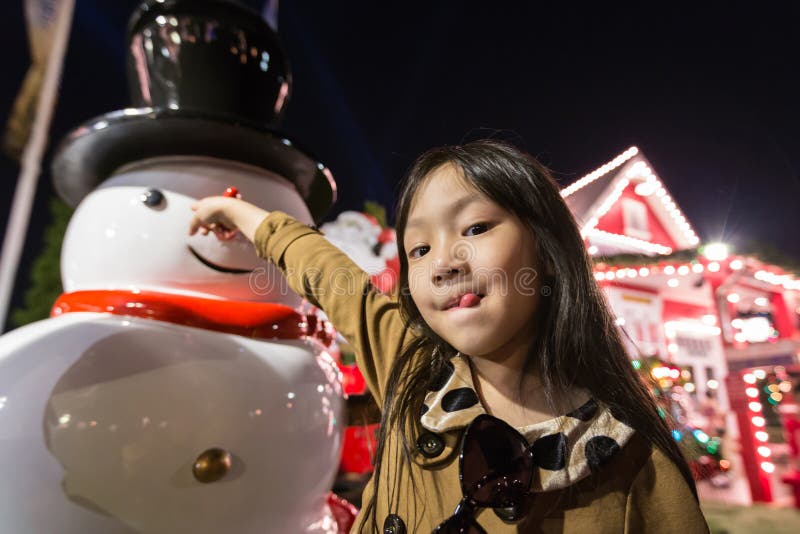 Posição asiática da menina e do boneco de neve no campo, opinião da noite no fundo da casa e árvore de Natal no year’s novos fotografia de stock royalty free Posição asiática da menina e do boneco de neve no campo, opinião da noite no fundo da casa e árvore de Natal no year’s novos fotografia de stock royalty free