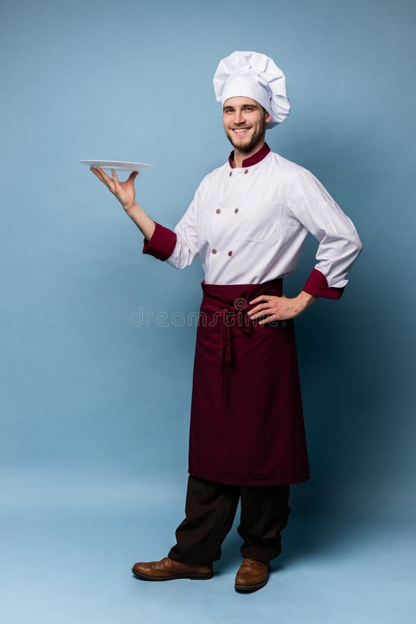 Posição masculina feliz do cozinheiro do cozinheiro chefe com a placa isolada em claro - fundo azul imagem de stock Posição masculina feliz do cozinheiro do cozinheiro chefe com a placa isolada em claro - fundo azul imagem de stock