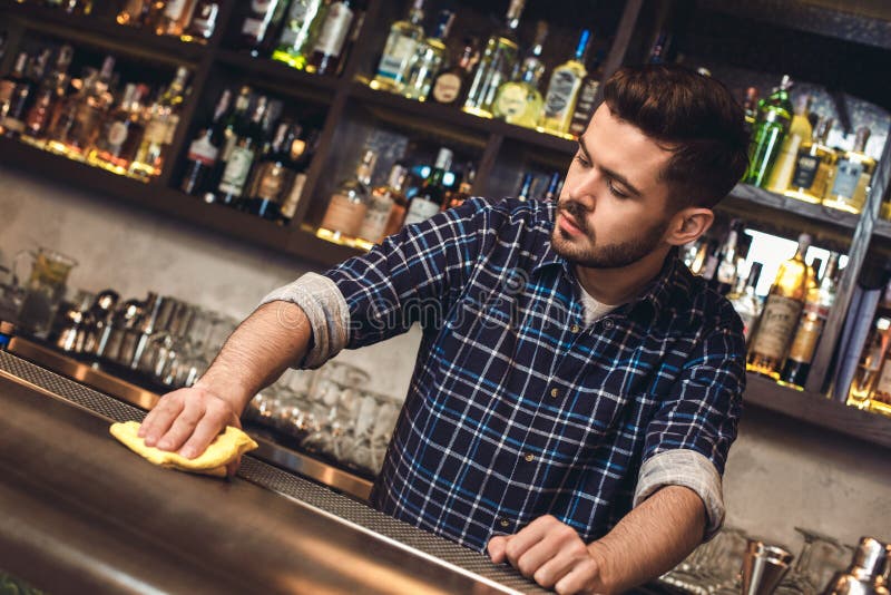 Posição nova do barman no contador da barra que limpa a superfície concentrada imagem de stock Posição nova do barman no contador da barra que limpa a superfície concentrada imagem de stock