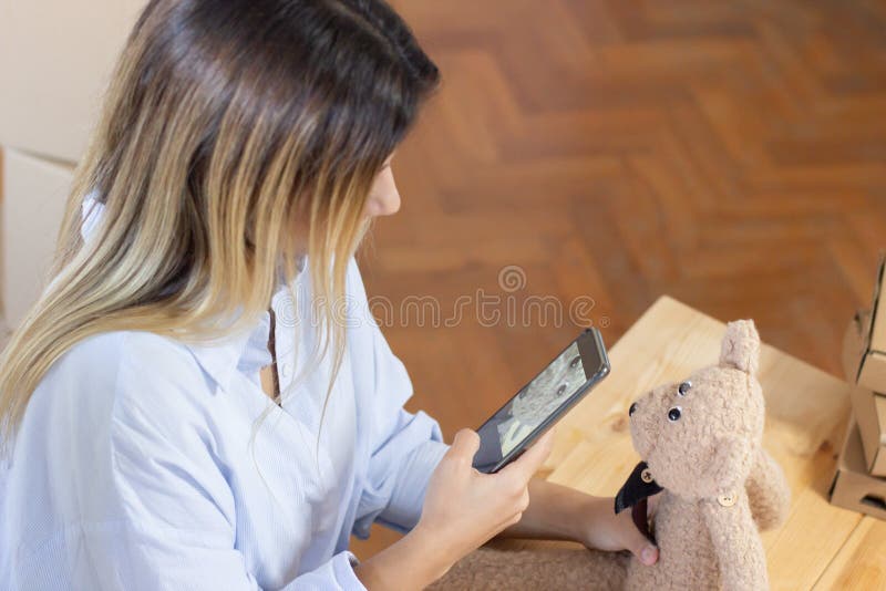 Postal worker photographing toy before dispatch. Young woman in blue shirt sitting at table and preparing product for packing. Shipping service concept