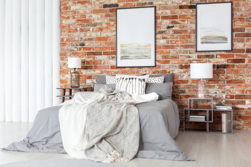 Posters on a bare, red brick wall, above a cozy bed with gray sheets in an industrial bedroom interior with natural light. Posters on a bare, red brick wall, above a cozy bed with gray sheets in an industrial bedroom interior with natural light