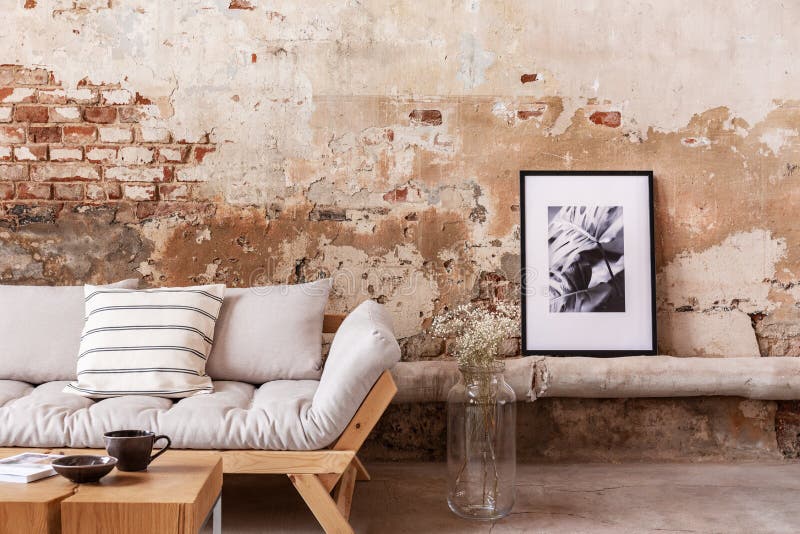 Poster and flowers next to grey wooden couch with pillows in flat interior with brick wall. Real photo