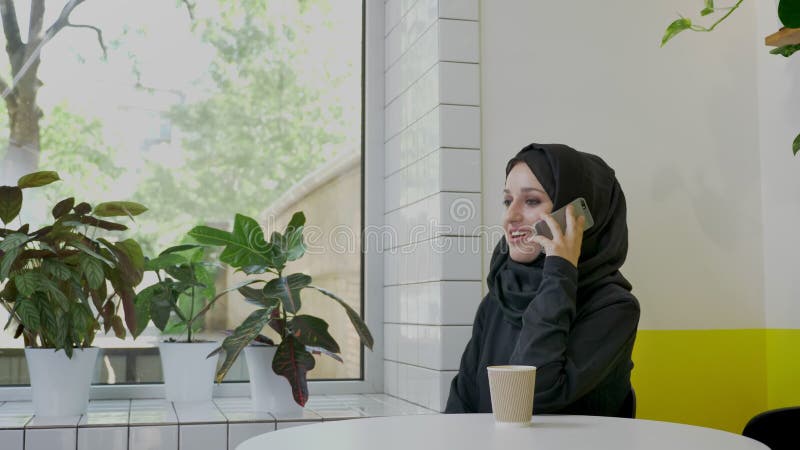 Potomstwo dosyć muzułmańska kobieta opowiada na telefonie i ono uśmiecha się w hijab, siedzi w kawiarni, rozochoconej