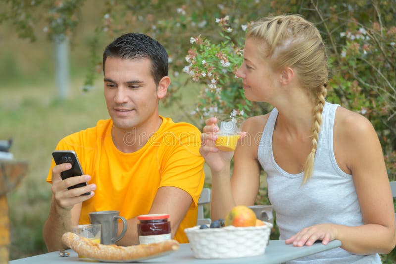 Potomstwo para sprawdza telefon podczas gdy mieć śniadanie outdoors obraz royalty free Potomstwo para sprawdza telefon podczas gdy mieć śniadanie outdoors obraz royalty free