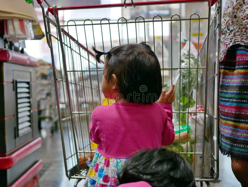 Pouco bebê asiático que ajuda sua mãe a empurrar um carrinho de compras em um supermercado fotografia de stock Pouco bebê asiático que ajuda sua mãe a empurrar um carrinho de compras em um supermercado fotografia de stock