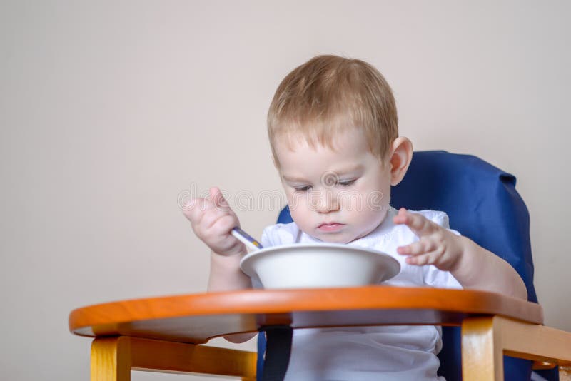 Pouco bebê que aprende comer em uma tabela que estuda uma placa e uma colher na cozinha imagens de stock royalty free Pouco bebê que aprende comer em uma tabela que estuda uma placa e uma colher na cozinha imagens de stock royalty free