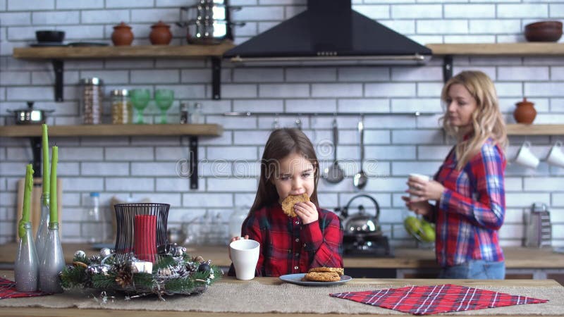 Pouco filha e mãe nova bonita nas camisas quadriculados que comem o café da manhã na cozinha junto A menina come