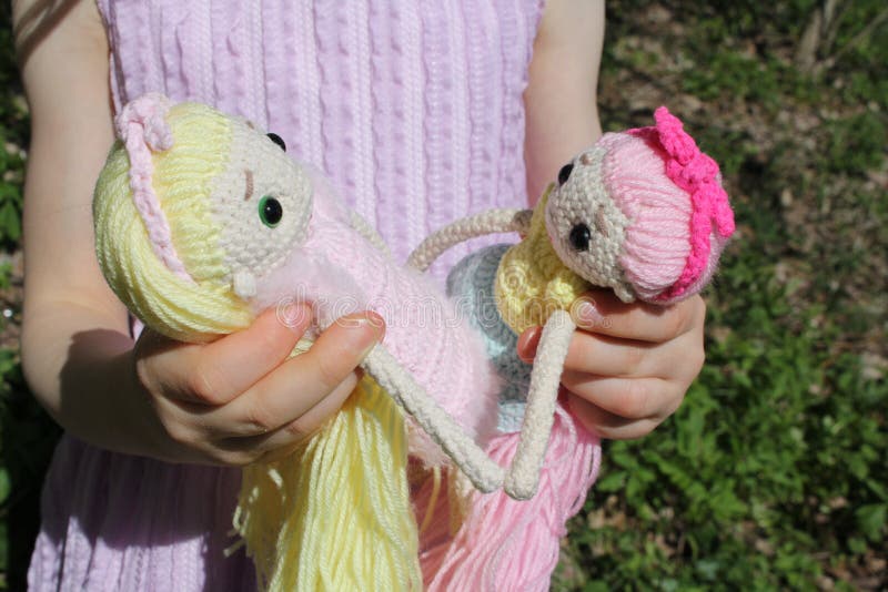 Dolls in the hands of an unrecognizable child in a pink dress. Knitted handmade dolls. Crochet. Hobby. A little girl holds in her hands 2 dolls with long hair in nature in a green field. High quality photo. Dolls in the hands of an unrecognizable child in a pink dress. Knitted handmade dolls. Crochet. Hobby. A little girl holds in her hands 2 dolls with long hair in nature in a green field. High quality photo