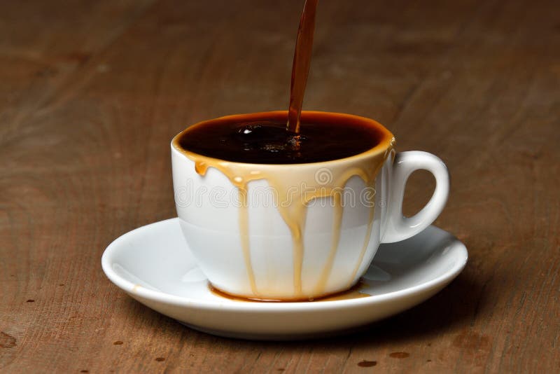 Pouring coffee into overflowed white porcelain mug with saucer. Cup with overflowing coffee. Full cup of coffee on wooden table