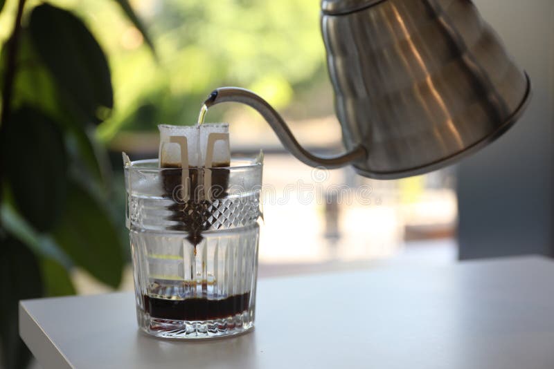 Pouring Hot Water into Glass with Drip Coffee Bag from Kettle on White Table Stock Photo Image