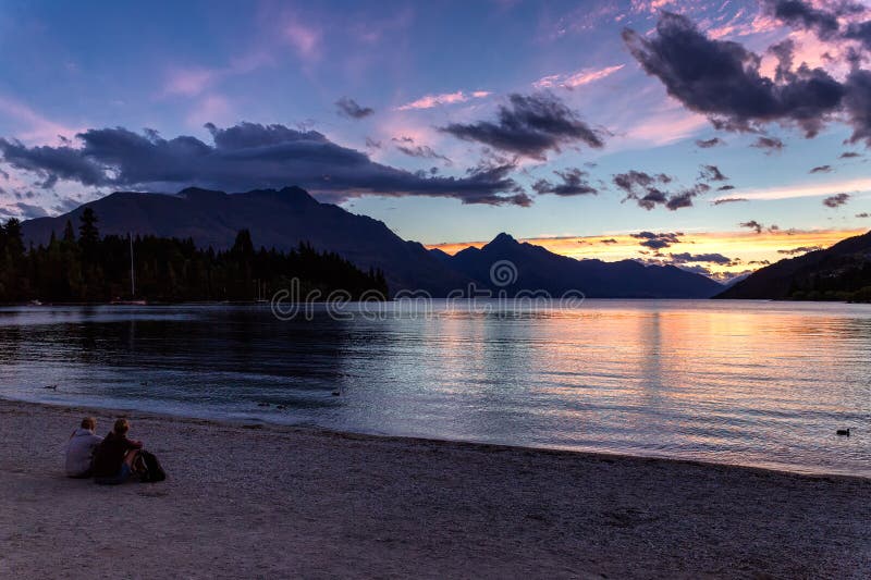 Povos que sentam-se pelo lago Wakatipu e pelo por do sol bonito de observação em Queenstown foto de stock royalty free Povos que sentam-se pelo lago Wakatipu e pelo por do sol bonito de observação em Queenstown foto de stock royalty free