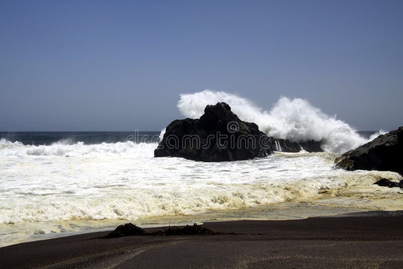 Powerfull waves crashing on a rock and splashing water in the air on remote black lava sand beach at Pacific coastline -