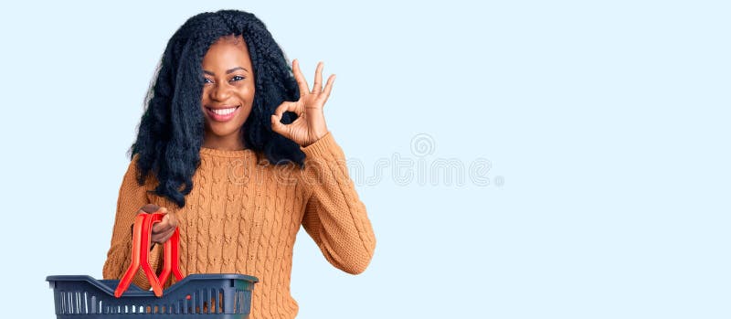 Beautiful african american woman holding supermarket shopping basket doing ok sign with fingers, smiling friendly gesturing excellent symbol. Beautiful african american woman holding supermarket shopping basket doing ok sign with fingers, smiling friendly gesturing excellent symbol