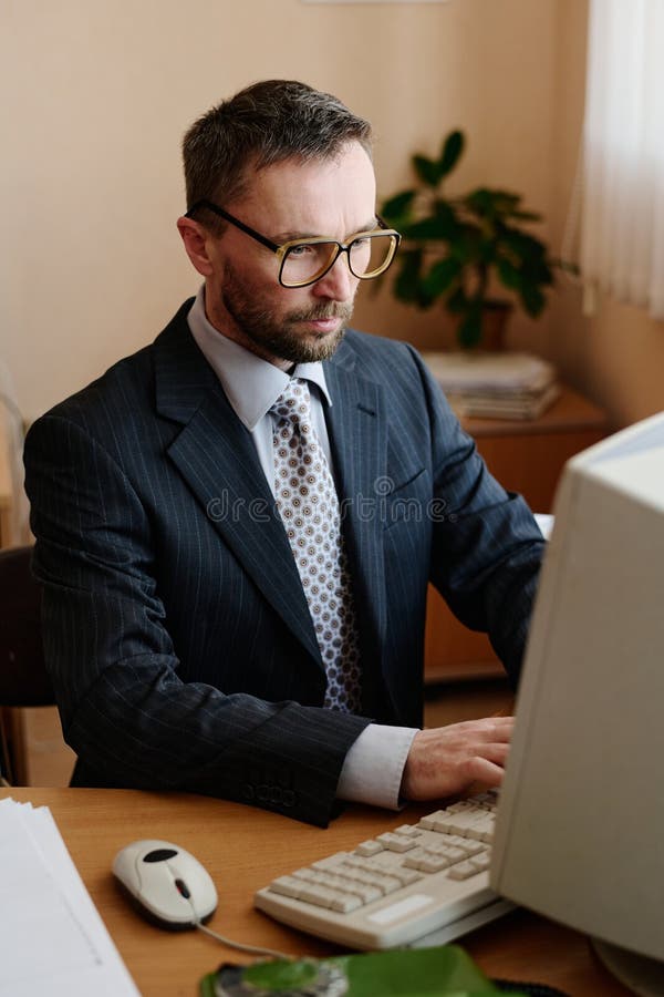 Pracujący profesjonalnie korzystający z komputera stacjonarnego w biurze. Biznesmen pracujący nad komputerem stacjonarnym w biurze nosi garnitur i okulary, koncentrując się na zadaniach z rośliną w tle, dodając naturalny wystrój
