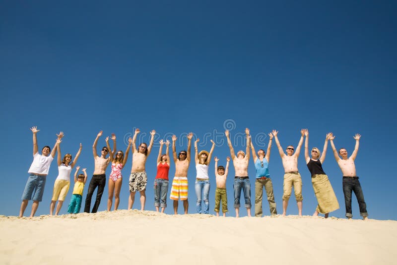 Image of many friends standing on sandy beach with their arms raised against blue sky. Image of many friends standing on sandy beach with their arms raised against blue sky