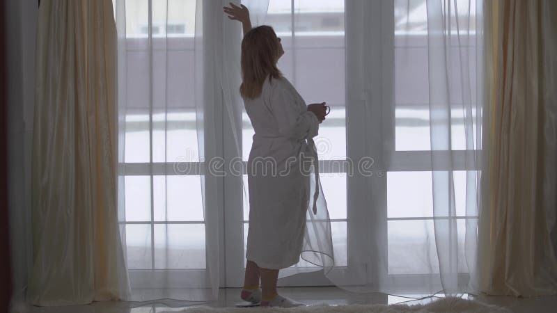 Pralles Mädchen im weißen langen Bademantel geht zum großen Fenster vom Boden bis zur Decke mit Tasse Kaffee oder Tee Damengerade