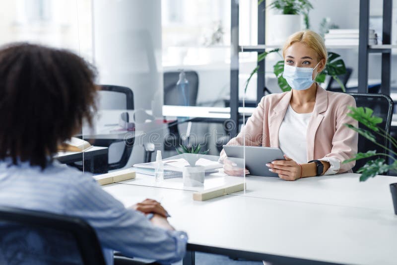 Talking to boss or interviewing during coronavirus epidemic. Young woman in protective mask with tablet communicates with african american lady through protective glass in modern office interior, empty space. Talking to boss or interviewing during coronavirus epidemic. Young woman in protective mask with tablet communicates with african american lady through protective glass in modern office interior, empty space