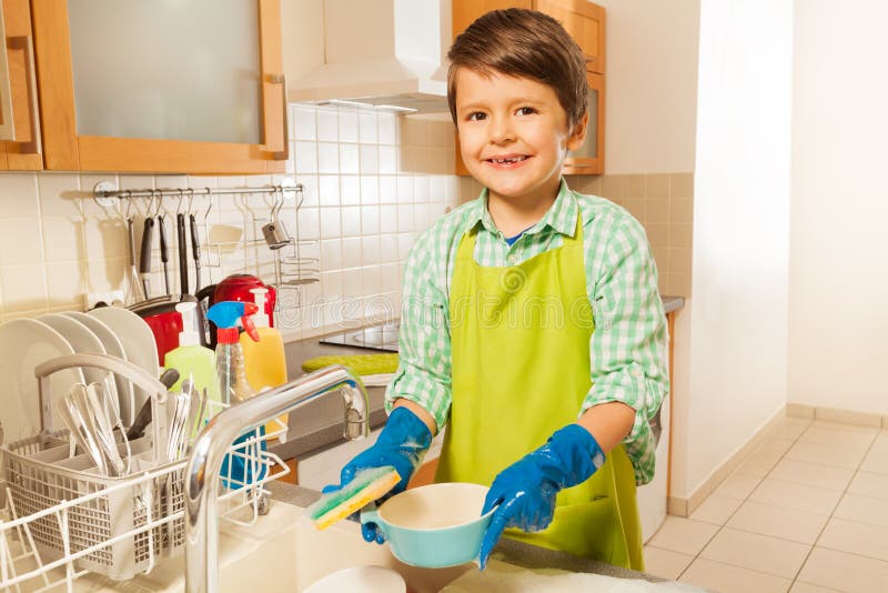 Lifestyle portrait of a boy washing dishes in the kitchen sink smiling to camera and holding mop with soap. Lifestyle portrait of a boy washing dishes in the kitchen sink smiling to camera and holding mop with soap