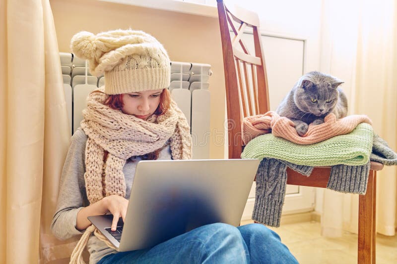 Preadolescente con sombrero de bufanda usando laptop calentando cerca de radiador de calefacción con gato gris mascota foto de archivo