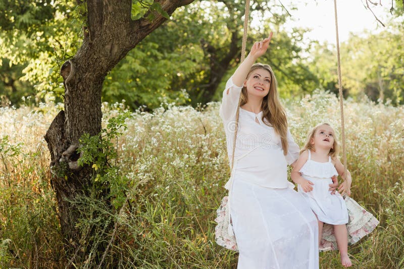 Pregnant beautiful mother with little blonde girl in a white dress sitting on a swing, laughing, childhood, relaxation, serenity, stock photography Pregnant beautiful mother with little blonde girl in a white dress sitting on a swing, laughing, childhood, relaxation, serenity, stock photography