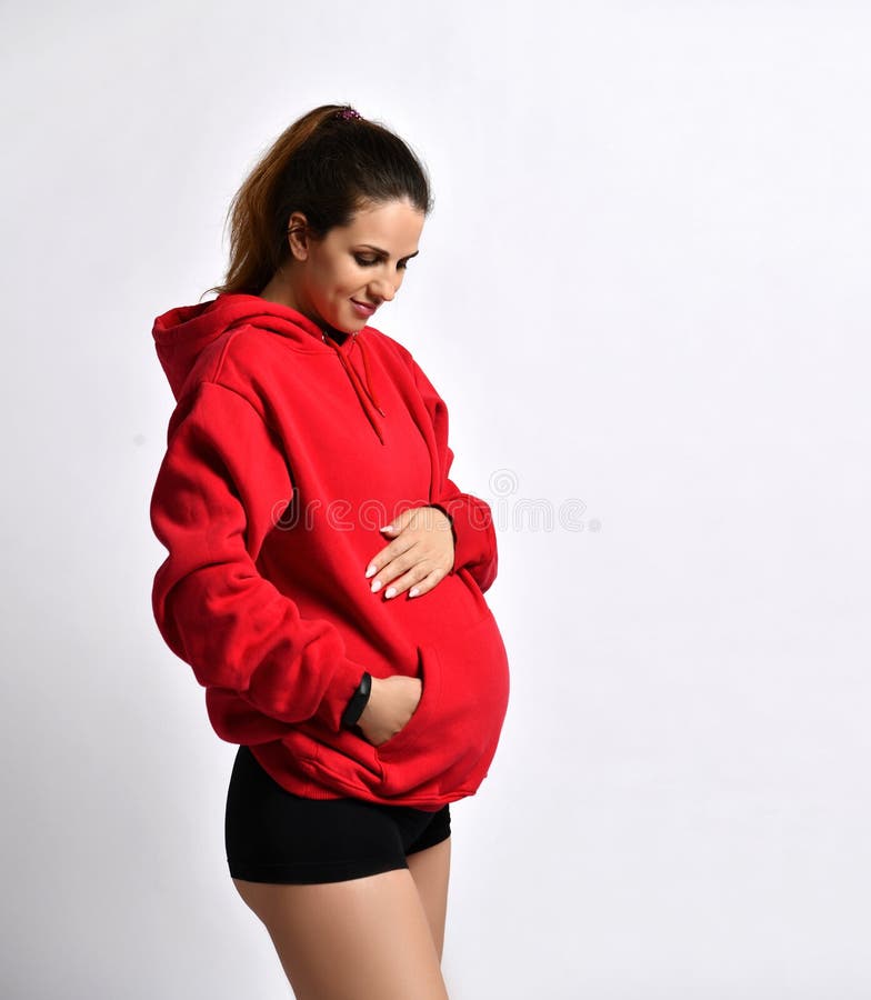 Pregnant female in black sport shorts, red hoody. She put hand on her belly, smiling, posing sideways isolated on white. Close up