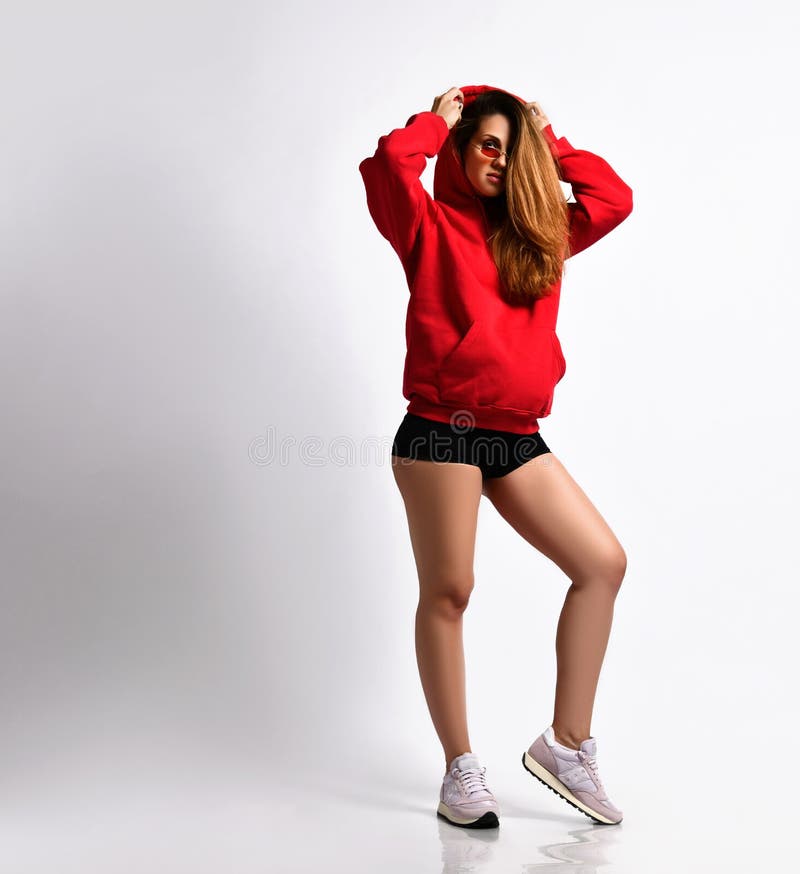 Pregnant lady in black shorts, red hoodie and sunglasses, pink sneakers. She is putting on a hood, isolated on white. Full-length