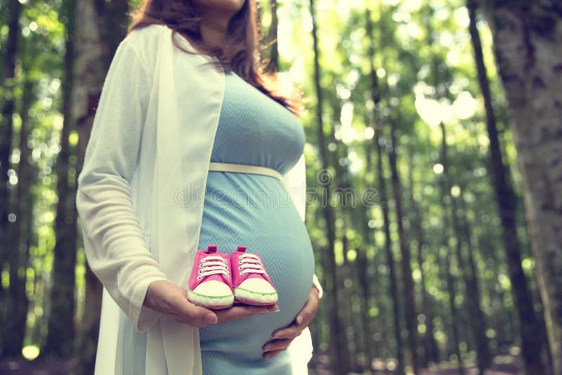 Pregnant woman holding a pair of pink sneakers toddler shoes while standing on the pine forest