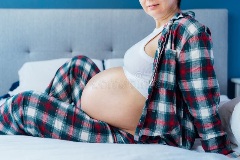 Pregnant woman sitting on bed, touching her belly, showcasing joy and anticipation. Expecting mother relaxing in pajama stock photo