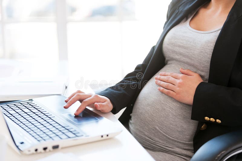 Cropped image of pregnant businesswoman typing something on laptop while sitting at her working place in office. Cropped image of pregnant businesswoman typing something on laptop while sitting at her working place in office