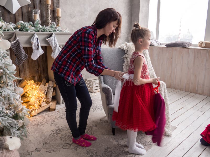 Preparando per il Natale: La mamma aiuta sua figlia messa su un vestito rosso per la palla di Natale fotografia stock Preparando per il Natale: La mamma aiuta sua figlia messa su un vestito rosso per la palla di Natale fotografia stock