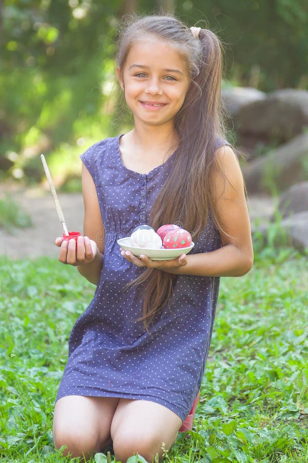 Cute smiling little girl sitting on a green grass and holding painted Easter eggs and paintbrus