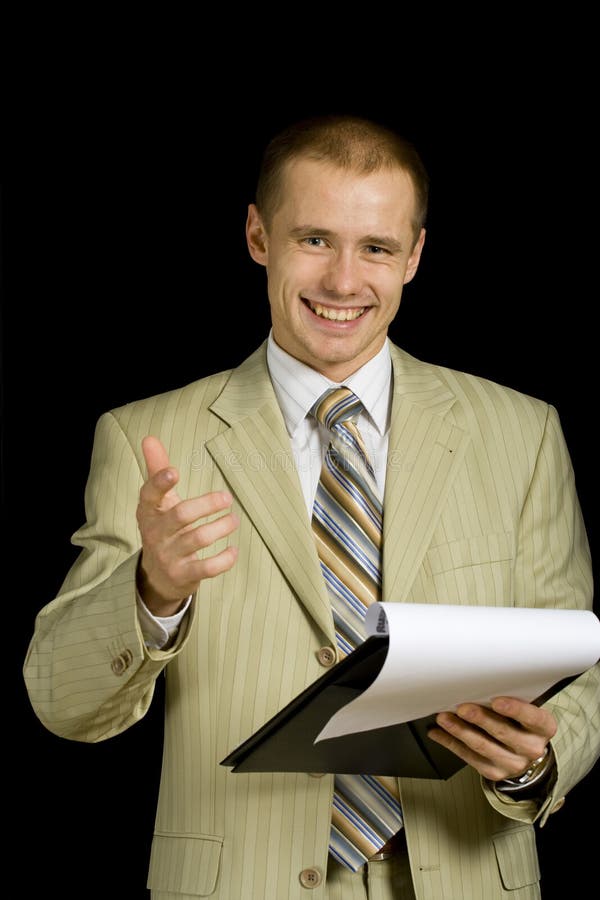 A studio portrait of a young businessman in a light colored business suit as he makes a presentation or sales pitch with gestures and holding a clipboard. Black or dark background. A studio portrait of a young businessman in a light colored business suit as he makes a presentation or sales pitch with gestures and holding a clipboard. Black or dark background.