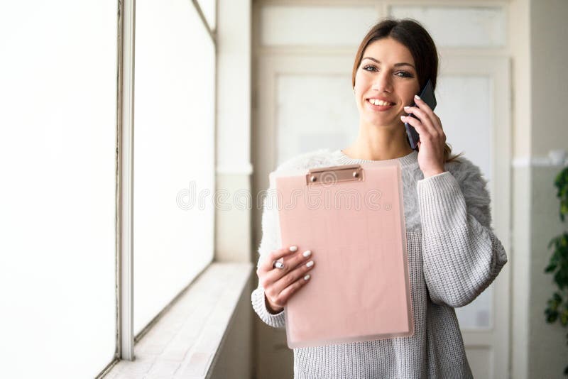 Presse-papiers de participation de jeune femme parlant au téléphone au travail photographie stock libre de droits Presse-papiers de participation de jeune femme parlant au téléphone au travail photographie stock libre de droits